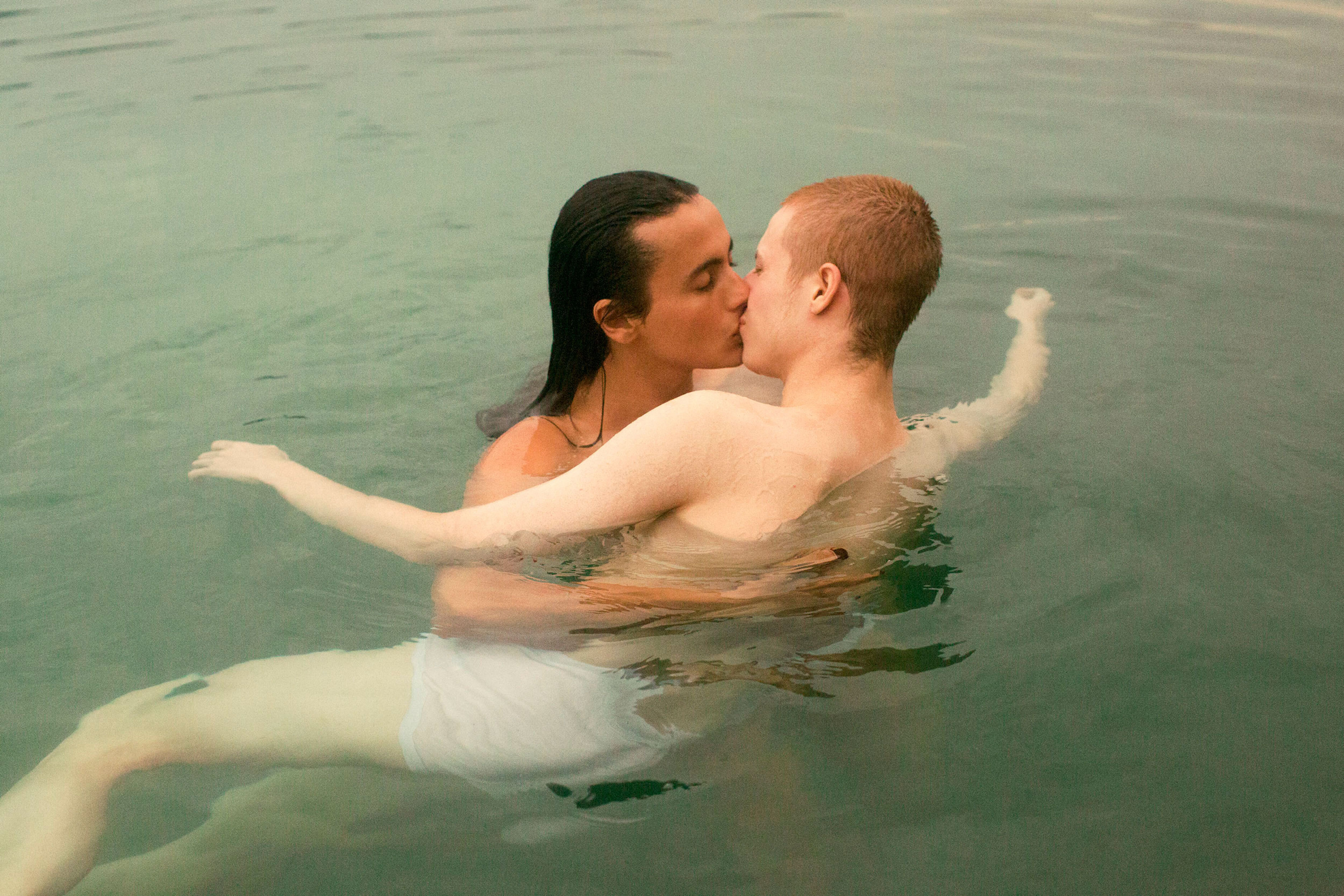 Two adult men embrace and kiss while standing waist-deep in pale green water outdoors on a day with soft, natural lighting.