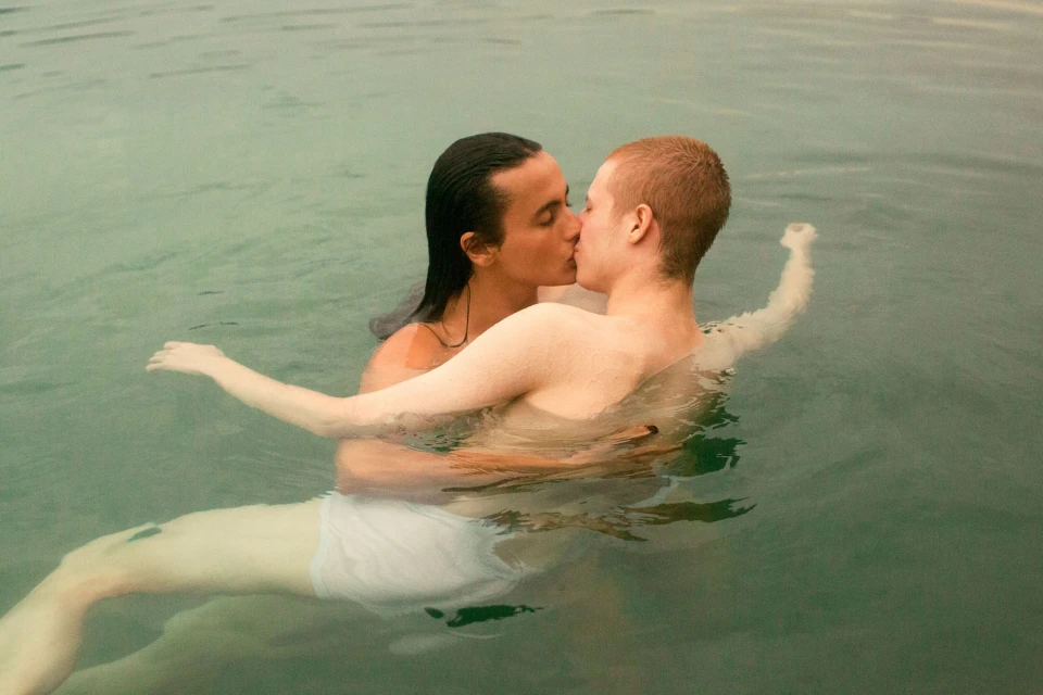 Two adult men embrace and kiss while standing waist-deep in pale green water outdoors on a day with soft, natural lighting.