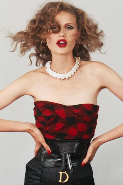 Blonde woman in red plaid top, black belt with D logo, and white necklace poses in a studio with her hands on her hips.