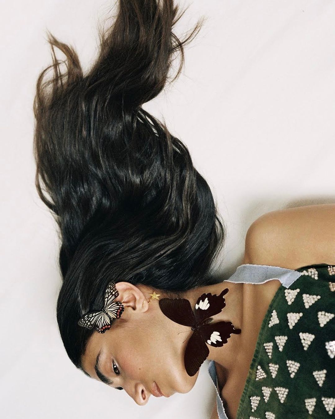 Brunette woman with butterfly earrings and green top poses on a white background in a beauty and fashion themed portrait.