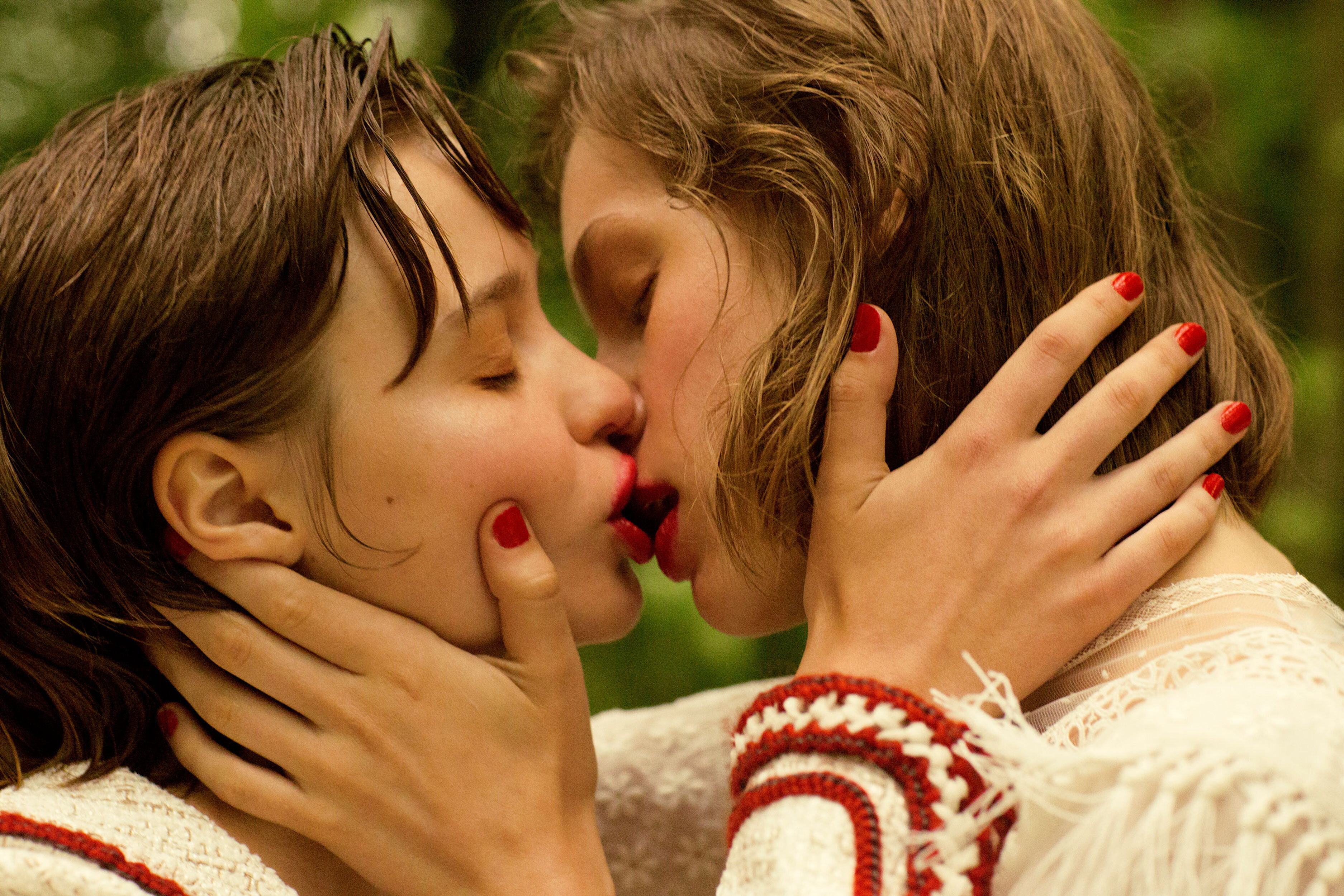 Close-up portrait of two fair-skinned adult females embracing and kissing in a lush green outdoor environment, emphasizing connection and intimacy.