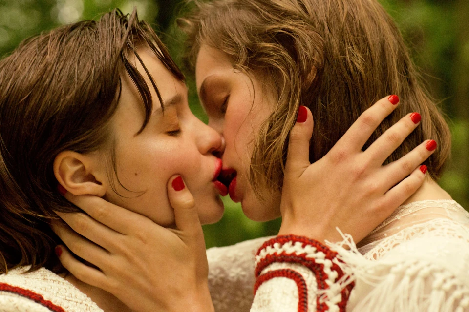 Close-up portrait of two fair-skinned adult females embracing and kissing in a lush green outdoor environment, emphasizing connection and intimacy.