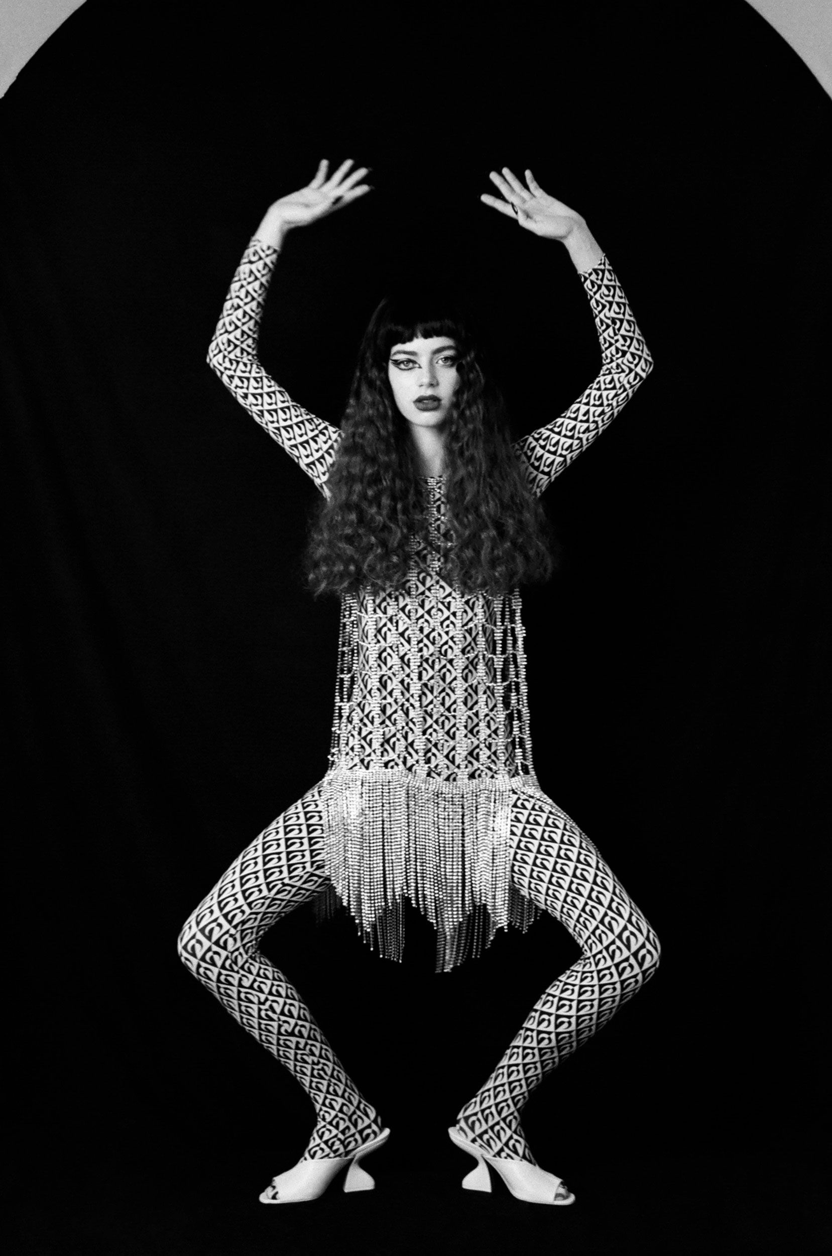 Black and white studio portrait of a woman in patterned long-sleeve top and leggings, fringe embellishments, bangs, and dark lipstick, arms raised.