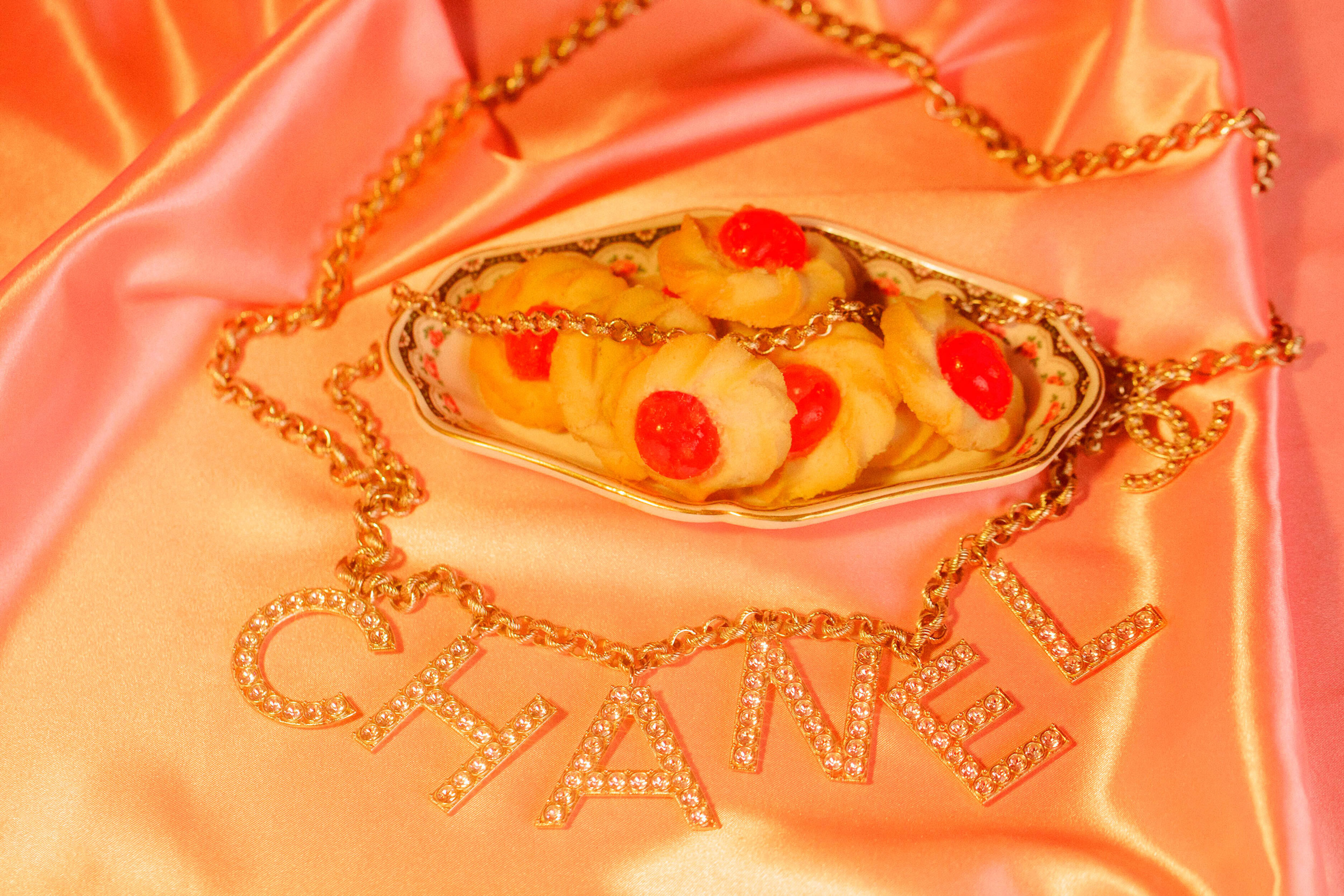 Still life of Chanel necklace, cookies with red cherry centers on a plate, and a pink satin background, evoking luxury and indulgence.