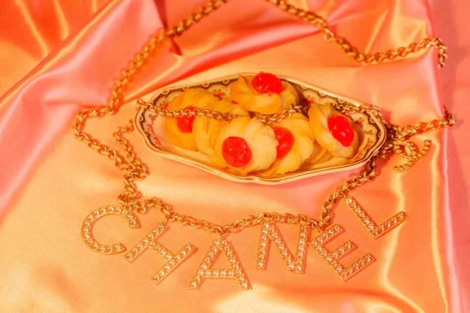 Still life of Chanel necklace, cookies with red cherry centers on a plate, and a pink satin background, evoking luxury and indulgence.