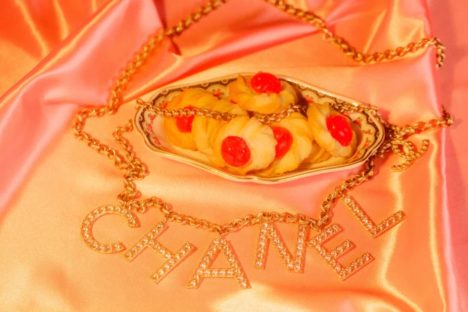 Still life of Chanel necklace, cookies with red cherry centers on a plate, and a pink satin background, evoking luxury and indulgence.