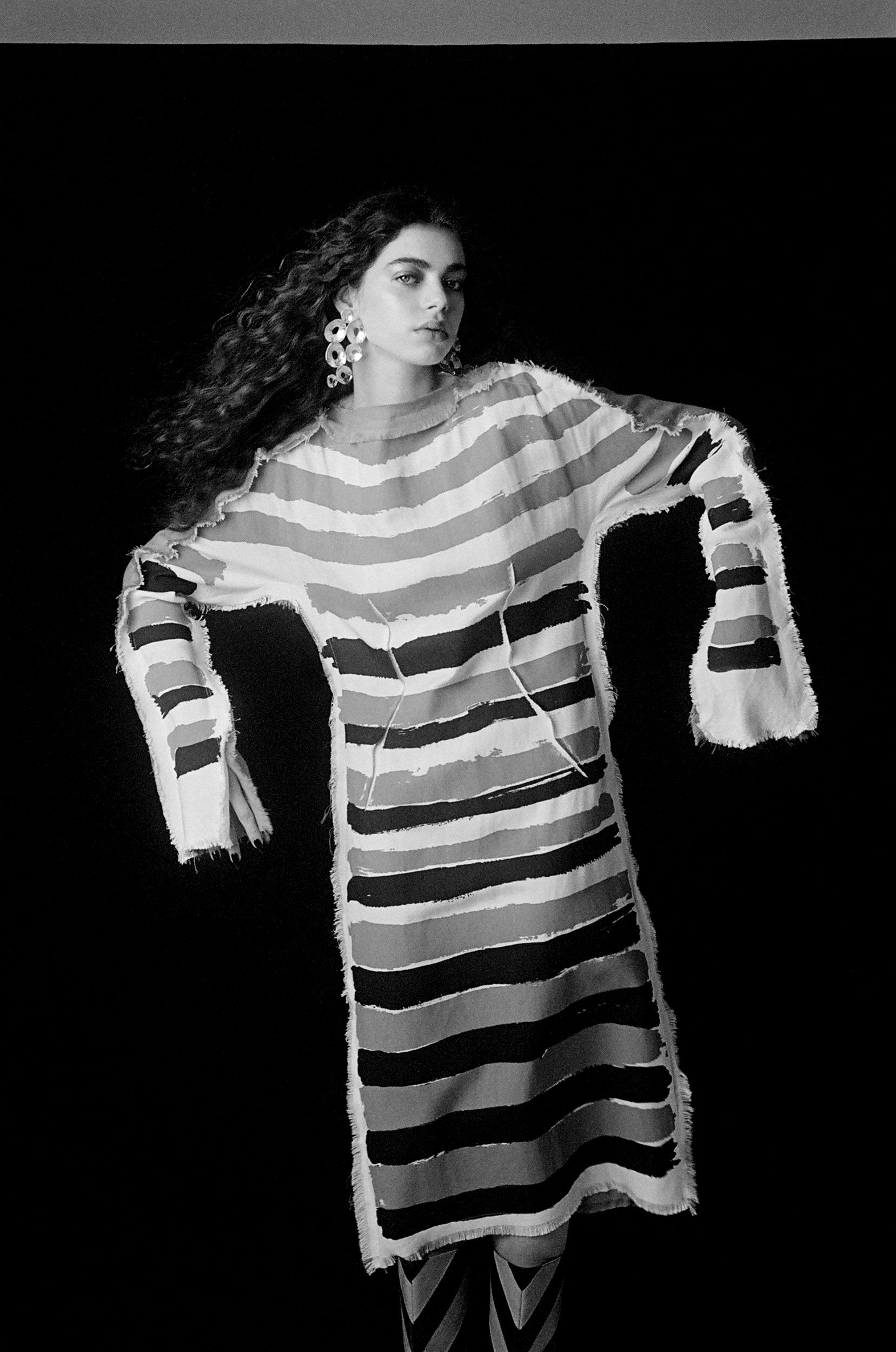 A woman with curly dark hair wears a striped vintage-inspired dress with large earrings in a studio setting with a black background.