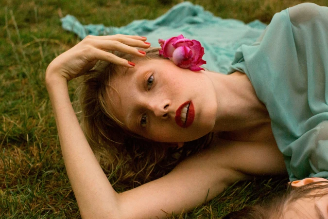 Woman lying on green grass wearing a turquoise dress with bright pink flowers tucked behind her ear.