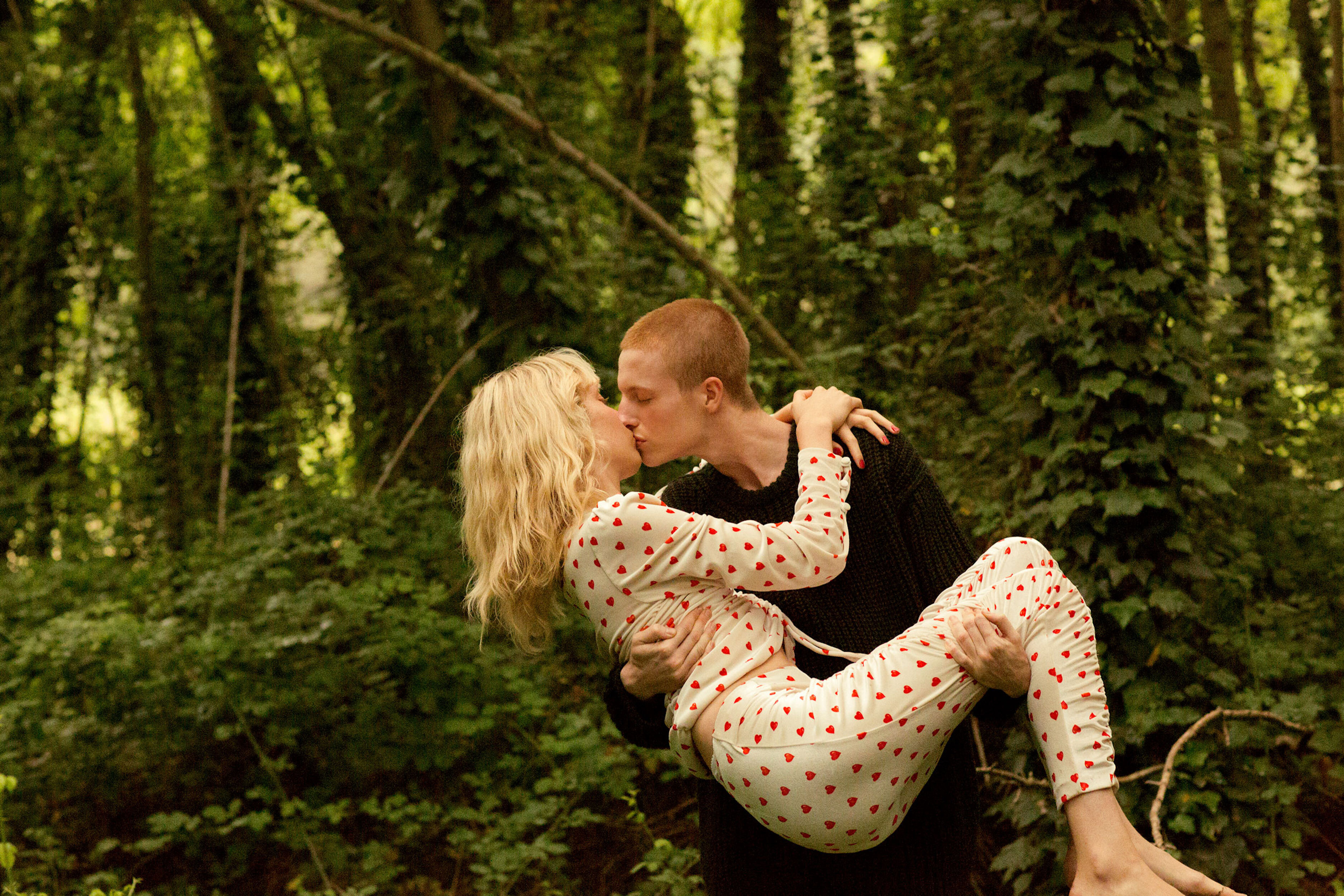 A couple kisses in a forest. The man holds the woman in his arms, with forest trees visible in the background, creating a romantic scene.
