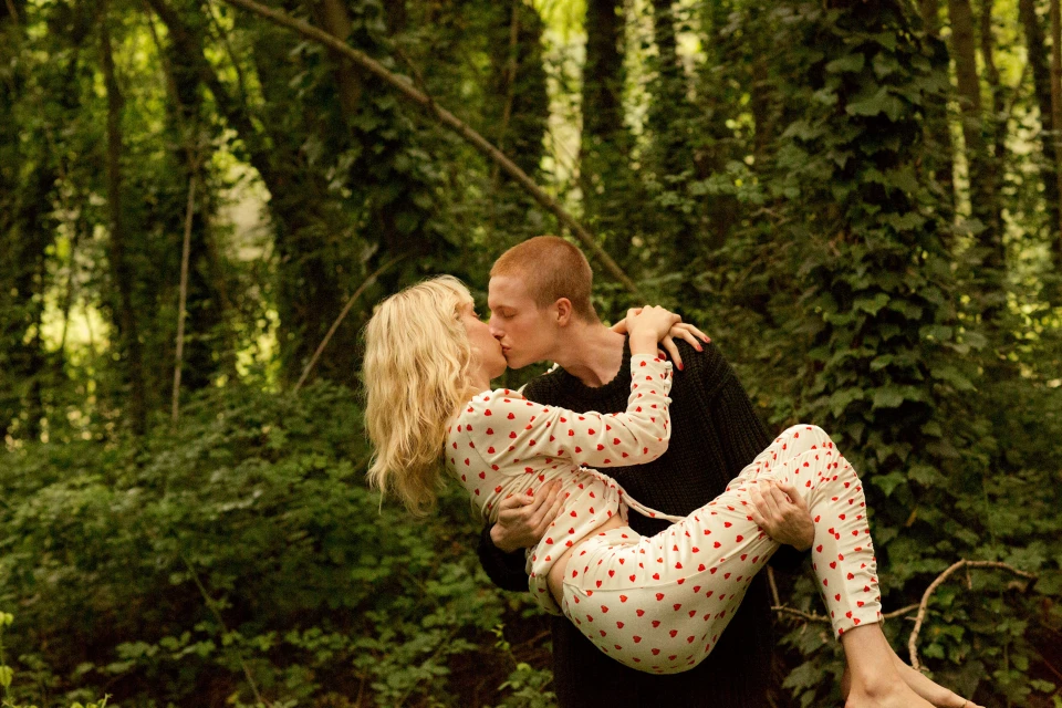 A couple kisses in a forest. The man holds the woman in his arms, with forest trees visible in the background, creating a romantic scene.