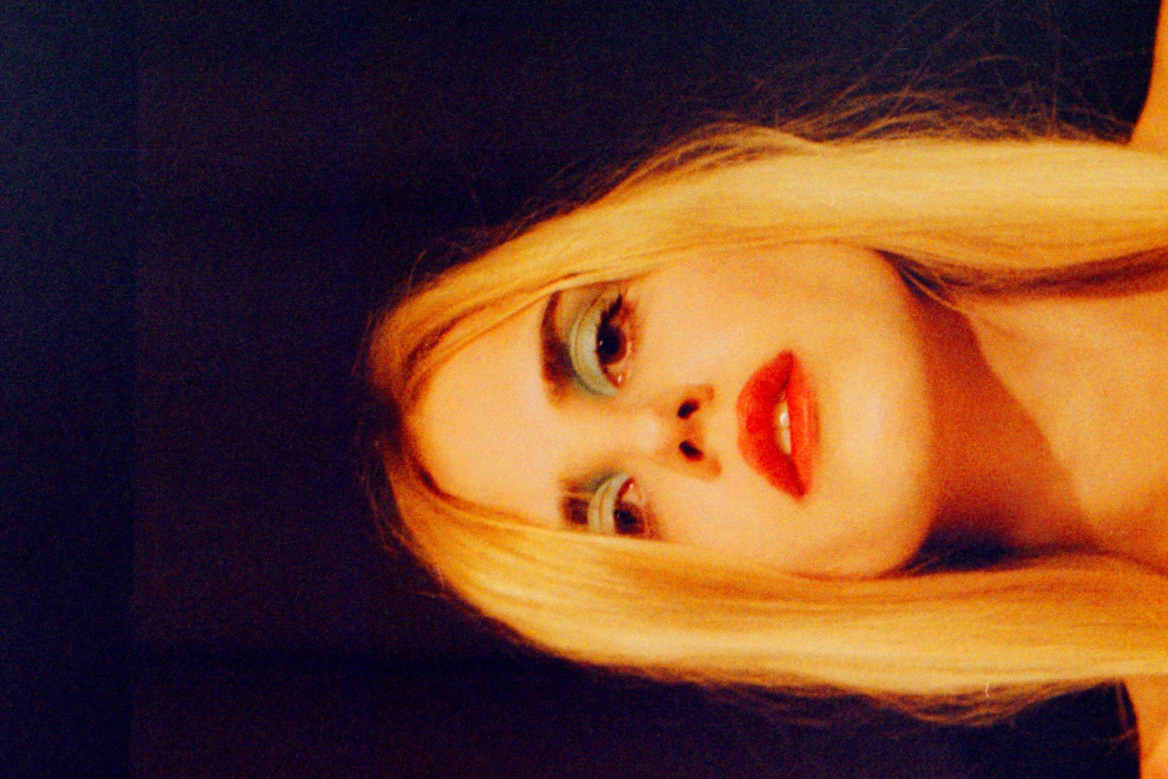 Adult female posing with blond hair, makeup and red lips against a dark background in a beauty headshot, shot with a shallow depth of field.
