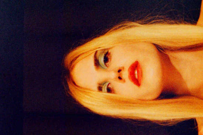 Adult female posing with blond hair, makeup and red lips against a dark background in a beauty headshot, shot with a shallow depth of field.