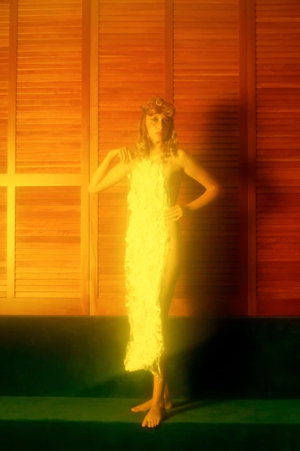 Adult female poses indoors, wearing a decorative yellow garment, hands on hips, with orange wood-paneled background and green velvet bench.