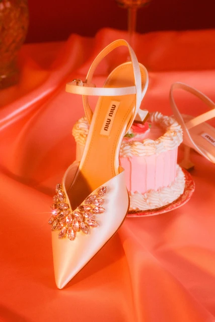Close-up studio shot of a satin high-heeled shoe adorned with rhinestones, alongside a pink layer cake on a silk coral surface.