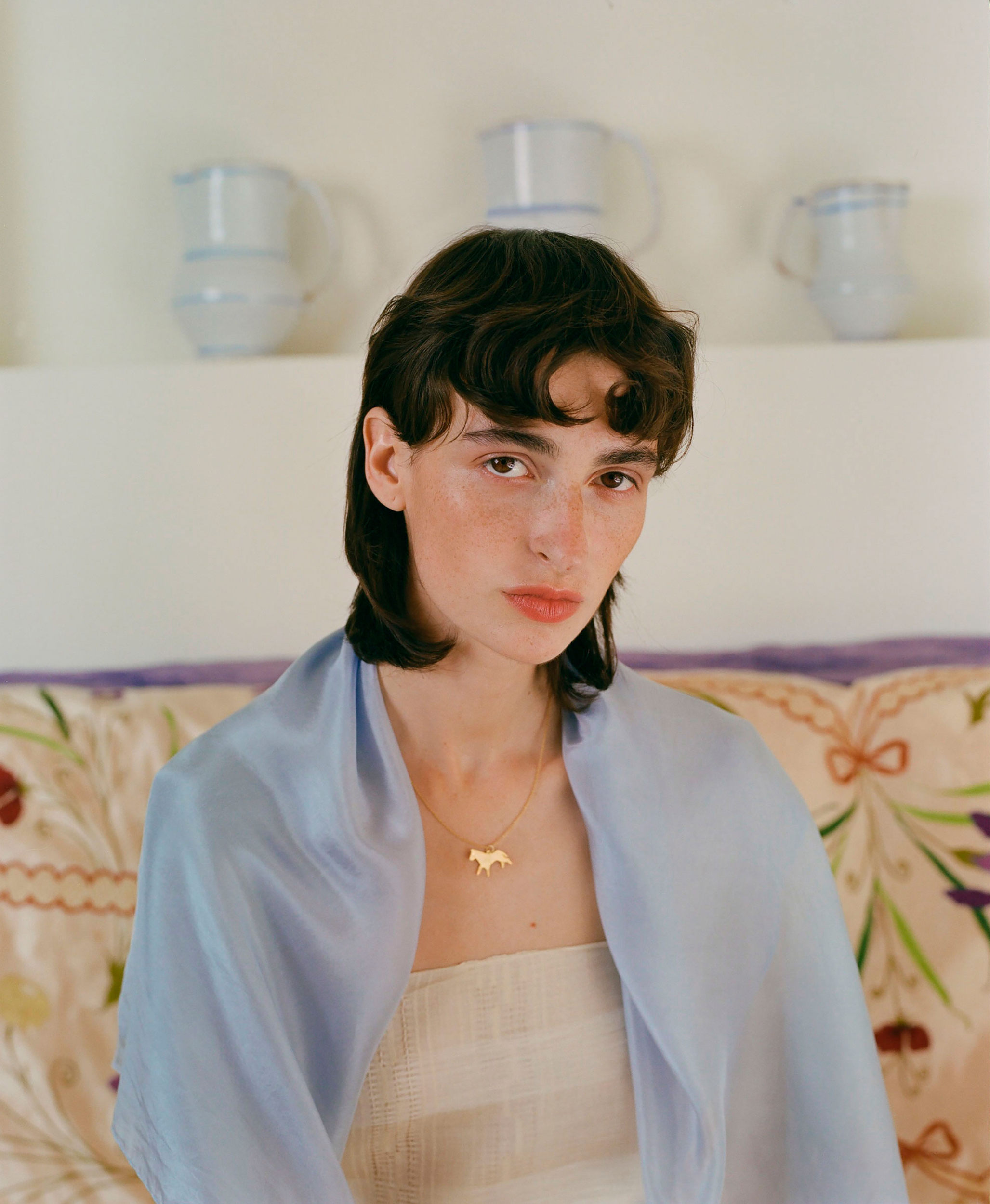 Close-up portrait of a woman with short dark hair wearing a pale blue scarf and gold necklace, posing in an indoor setting with white walls.