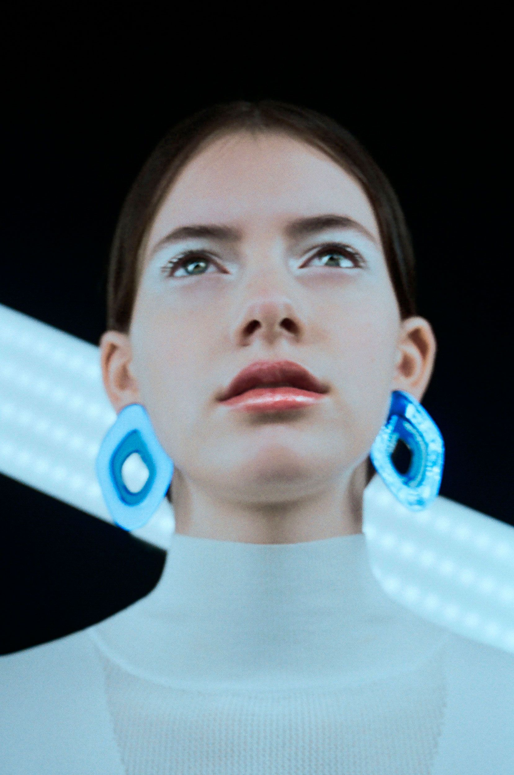 Portrait of a young woman with blue earrings and makeup, looking up against a black backdrop with white light.