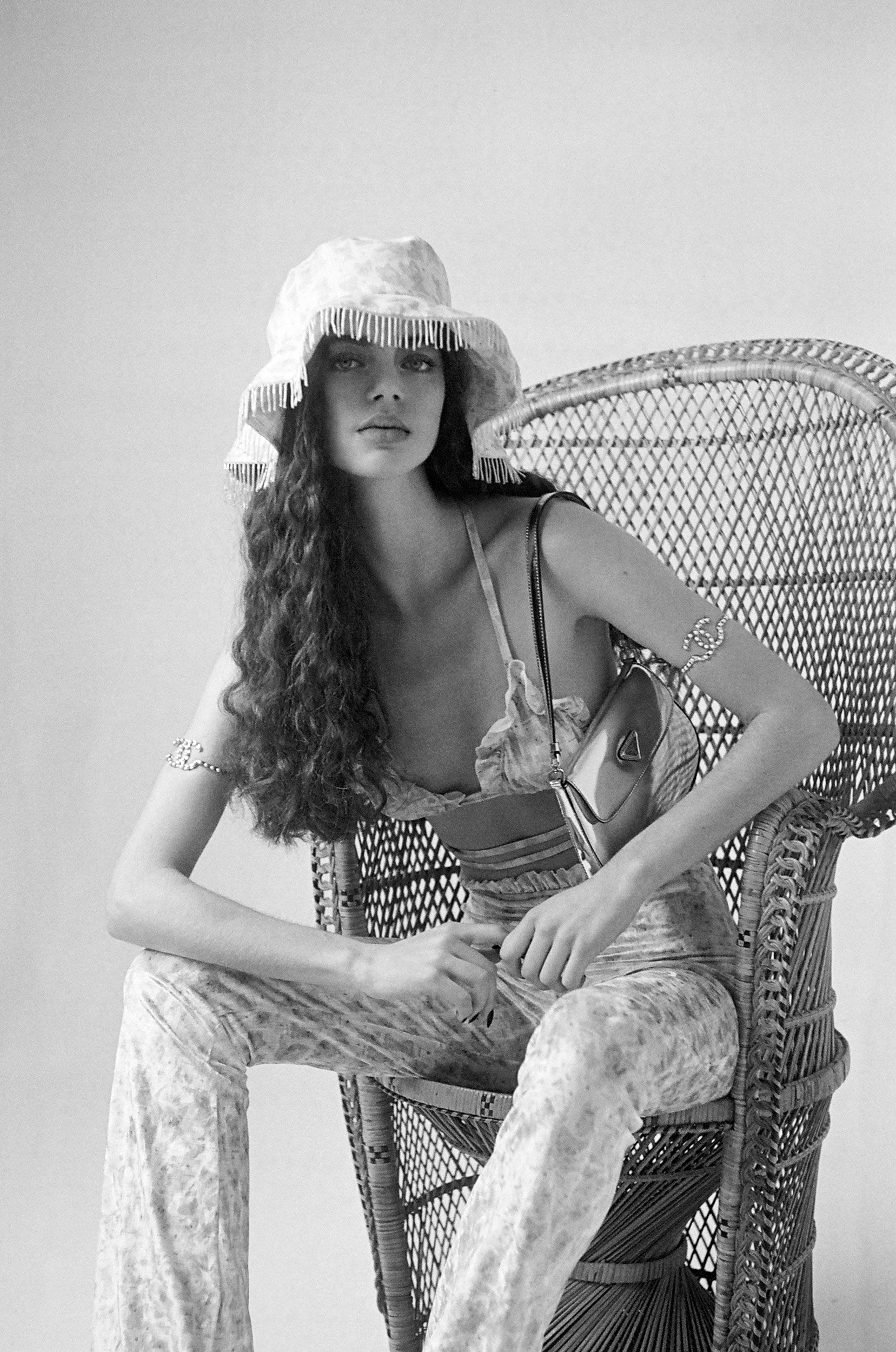 A woman in a floral-patterned outfit and hat poses in a wicker peacock chair in a studio setting with soft, monochromatic lighting.