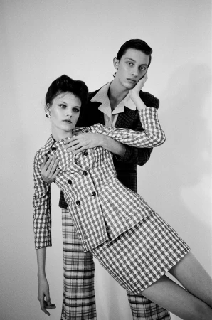 Black-and-white fashion studio portrait of a woman in a checked suit with a man behind her, hand on face, against a white background.