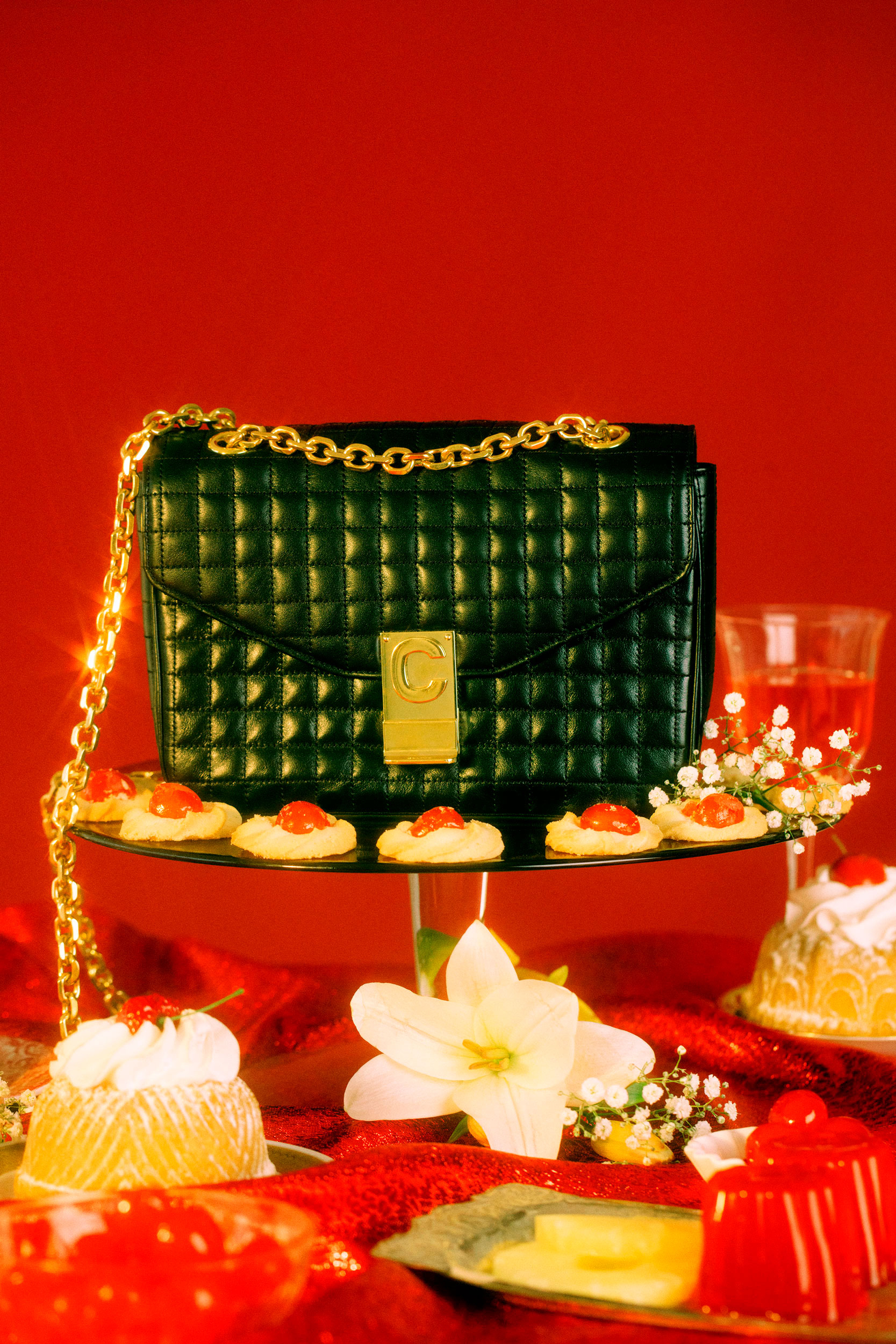 Elevated view of a black purse with a gold chain on a dessert table with jello, cookies, and flowers against a vibrant red backdrop.