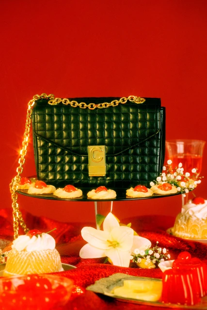 Elevated view of a black purse with a gold chain on a dessert table with jello, cookies, and flowers against a vibrant red backdrop.