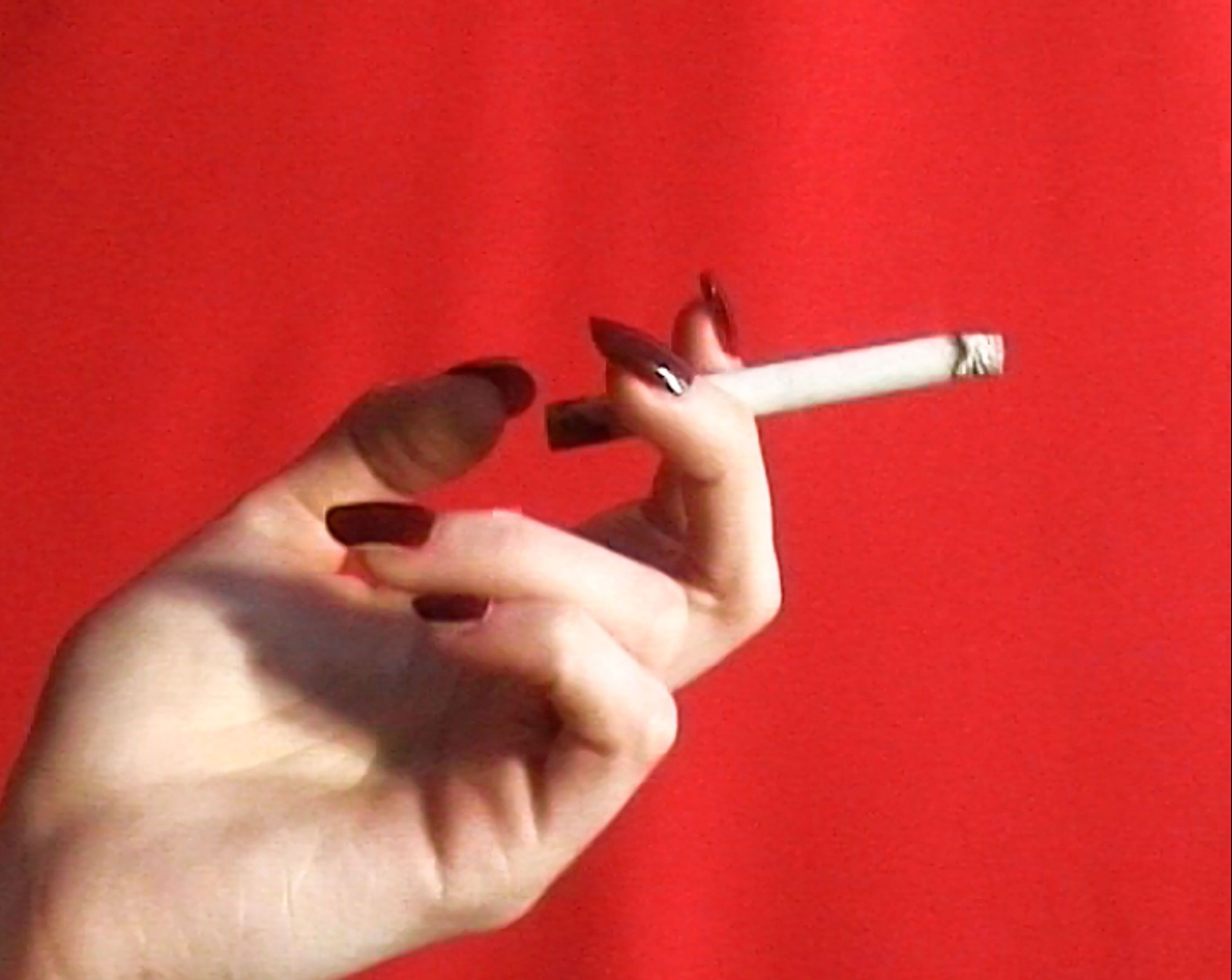 Close-up shot of a fair-skinned human hand holding a cigarette against a red background, showcasing long, dark-polished nails.