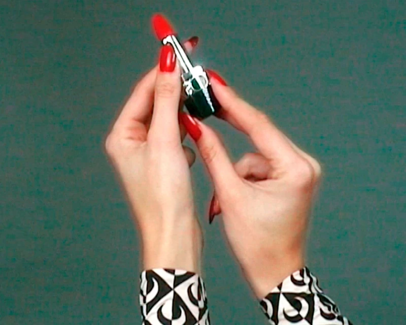 Hands holding red lipstick tube against a teal background. Woman with red nails and black-and-white patterned sleeve visible.