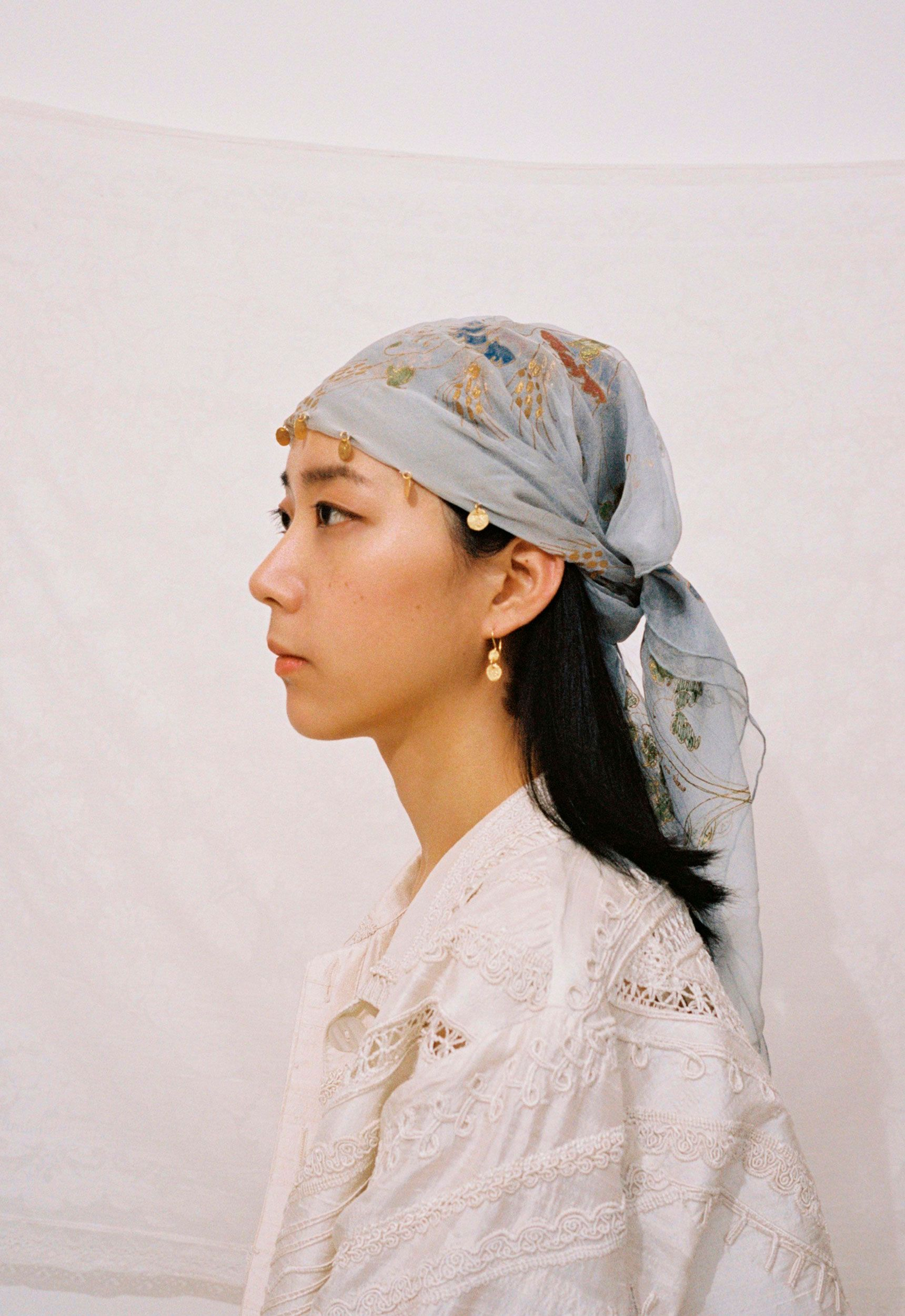 Portrait of a woman wearing a blue patterned headscarf and a white lace jacket, standing in front of a bright white backdrop.