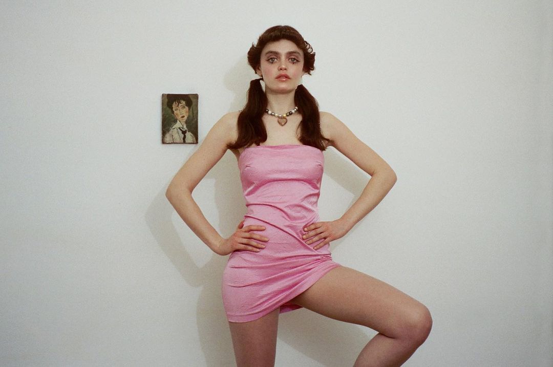 Young woman with pigtails poses in a pink dress in front of a white wall with an unframed portrait hanging on the wall.
