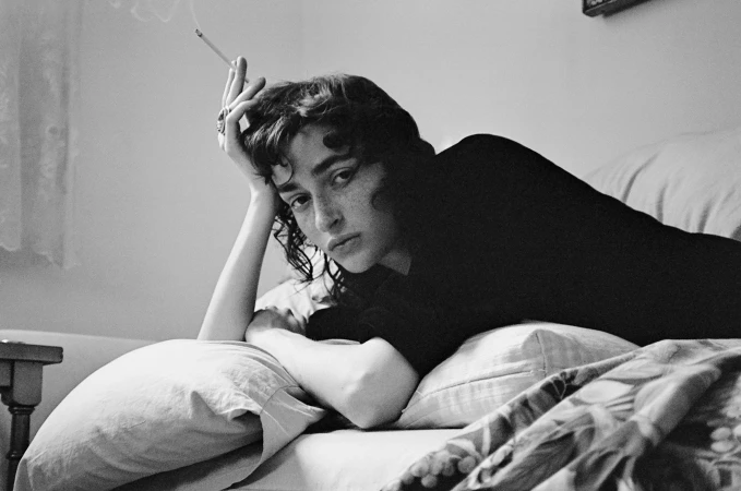 Monochrome portrait of a woman lying on a bed holding a lit cigarette while looking at the camera.
