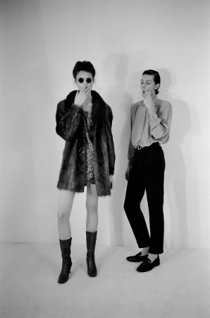 Two models smoking against a white backdrop, one wearing sunglasses and a fur coat.