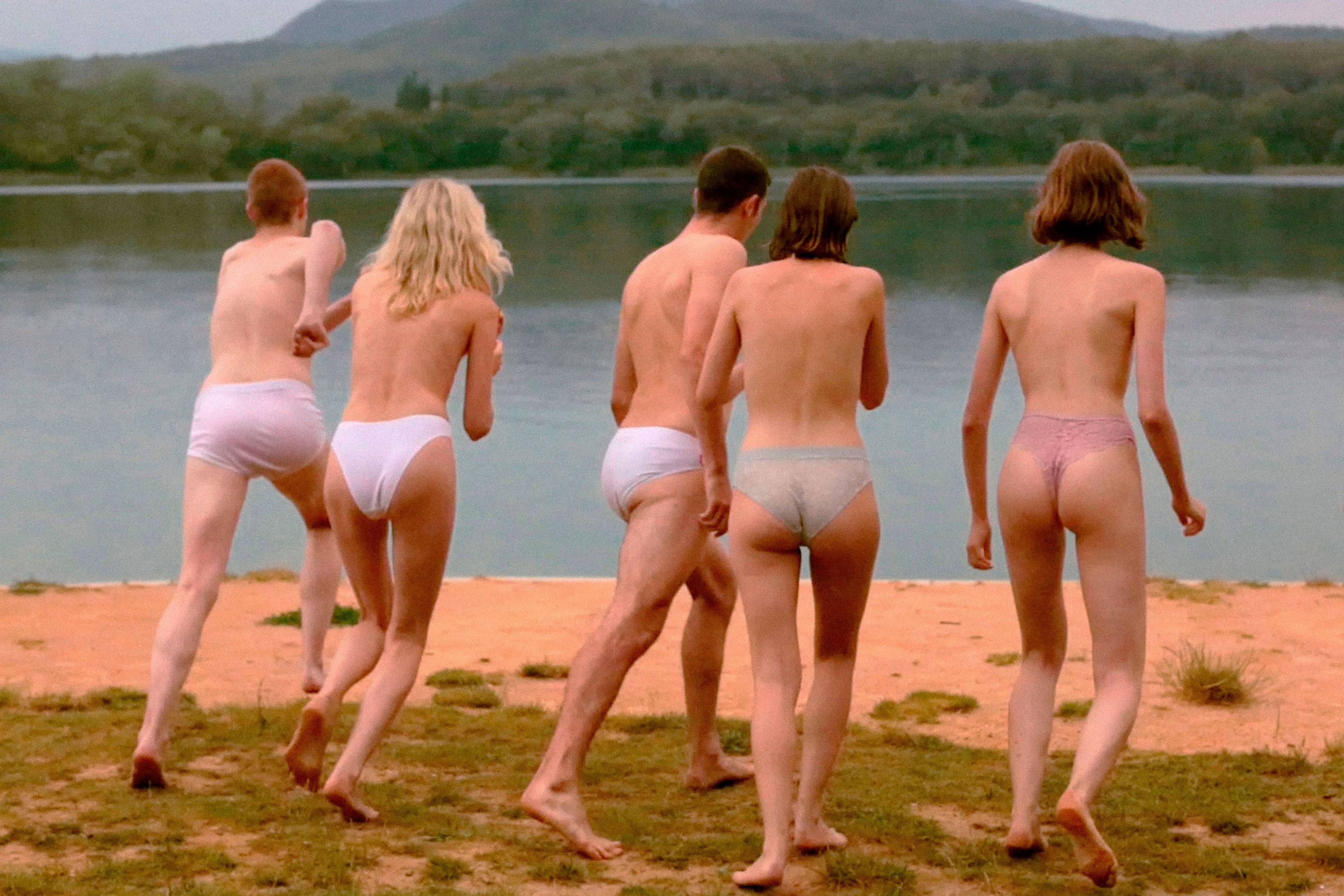 Group of young adult men and women in underwear, barefoot, standing by a lake surrounded by trees and hills, sunny weather, daylight.