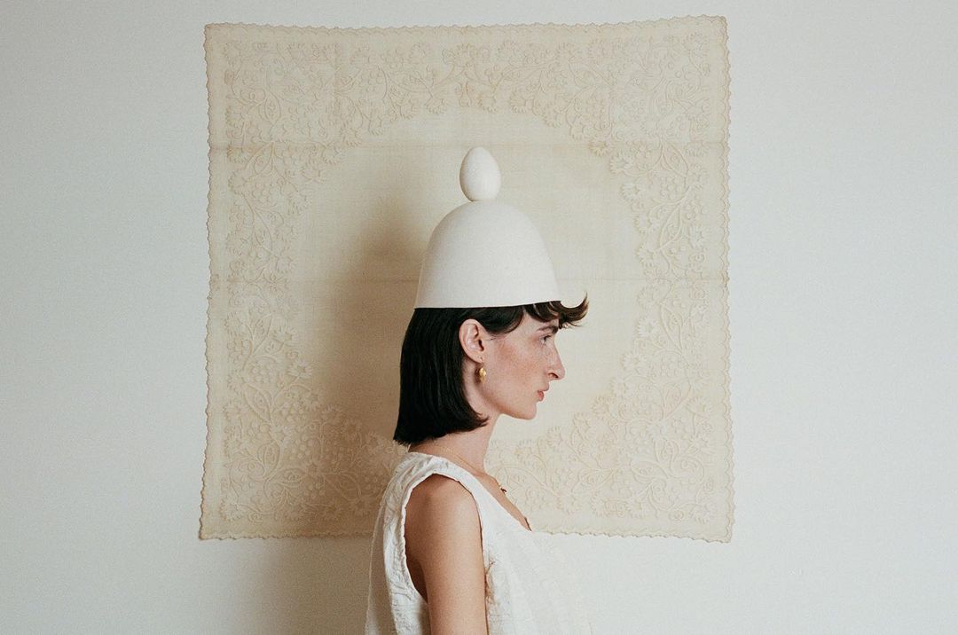 Profile of woman wearing white hat with egg atop against off-white lace and white wall. Earring and top visible.