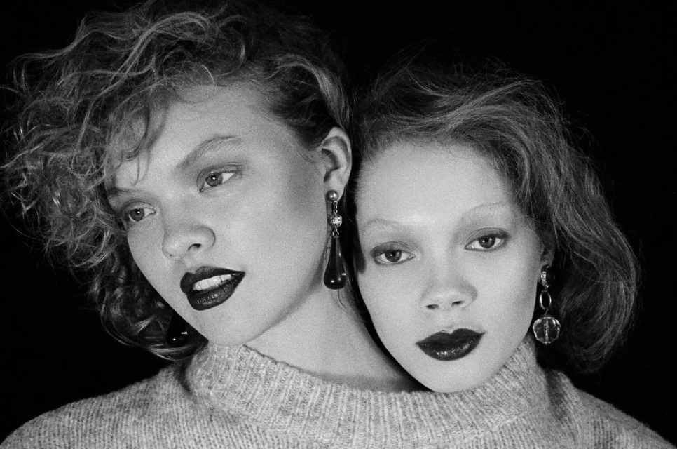 Black and white portrait of two young women with curly hair, dark lipstick, and teardrop earrings, wearing a grey wool sweater.