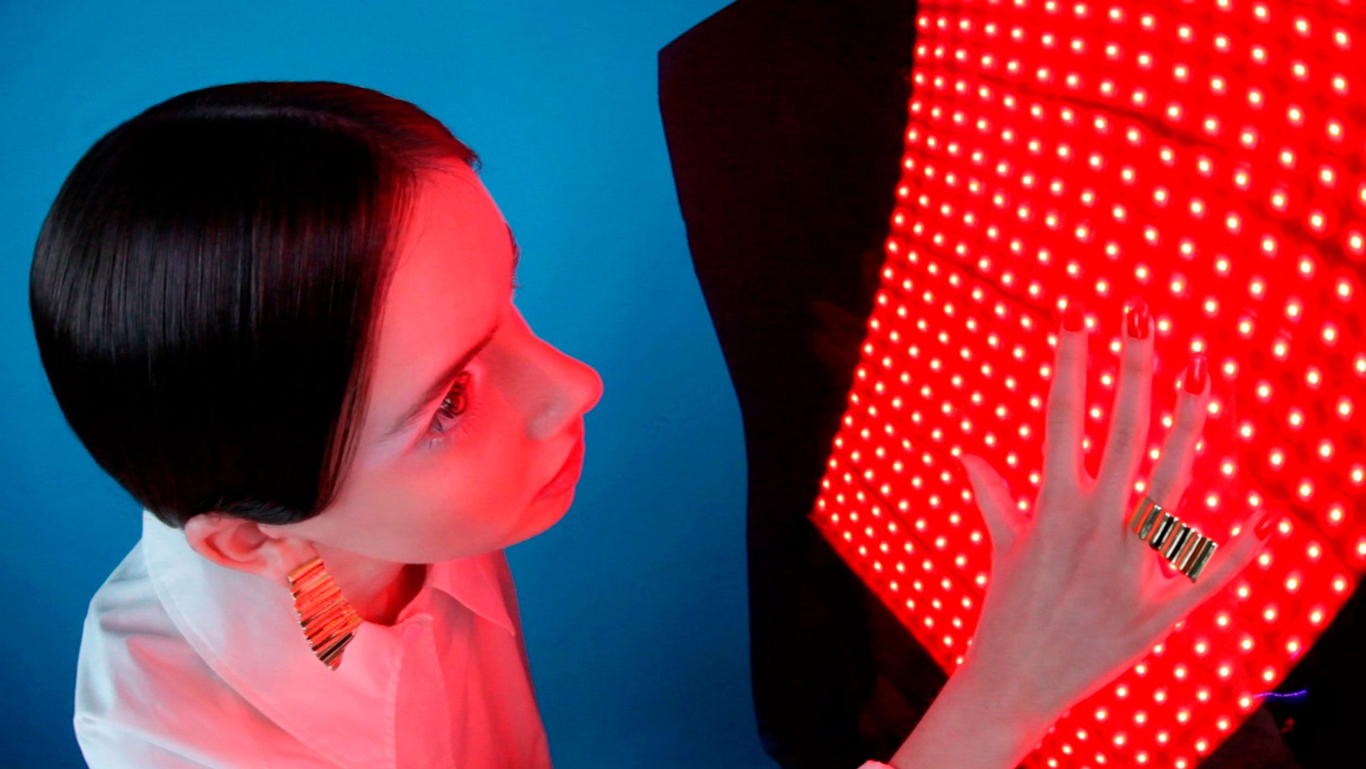 A young woman with dark hair, stylish jewelry, and painted red nails interacts with a modern red light therapy device in a studio setting.