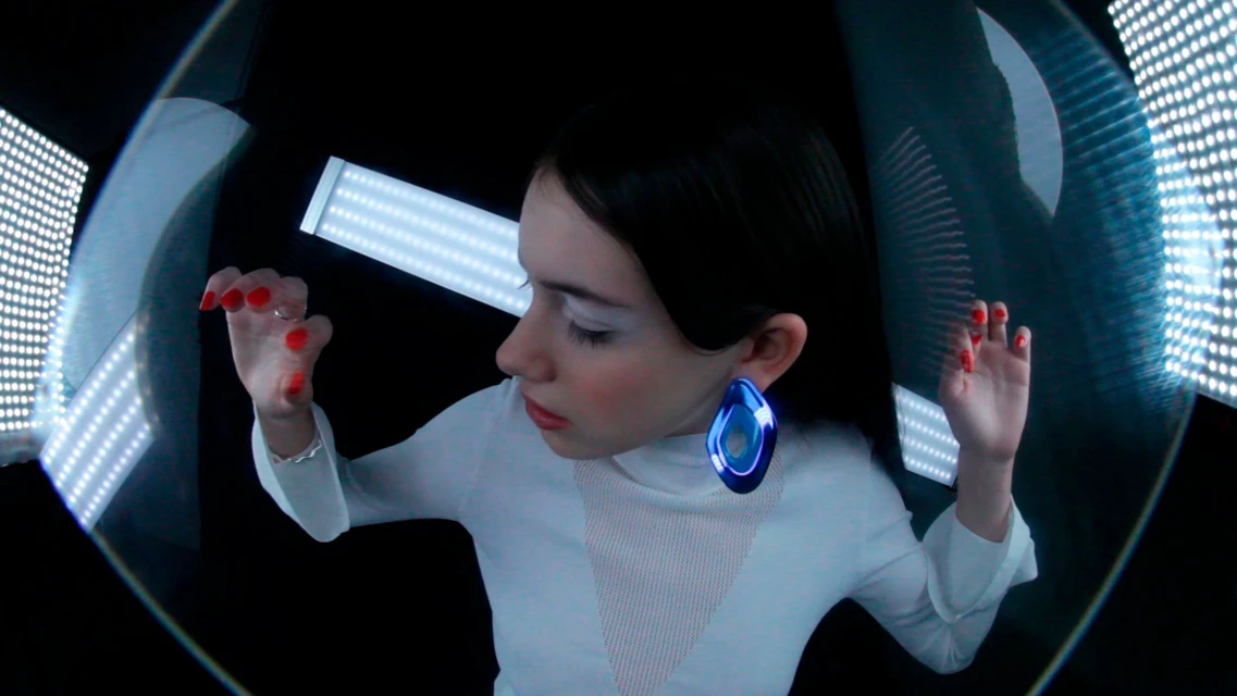 Artistic studio shot of a woman with face makeup and a large blue earring, inside a light box with red nail polish on her fingers.