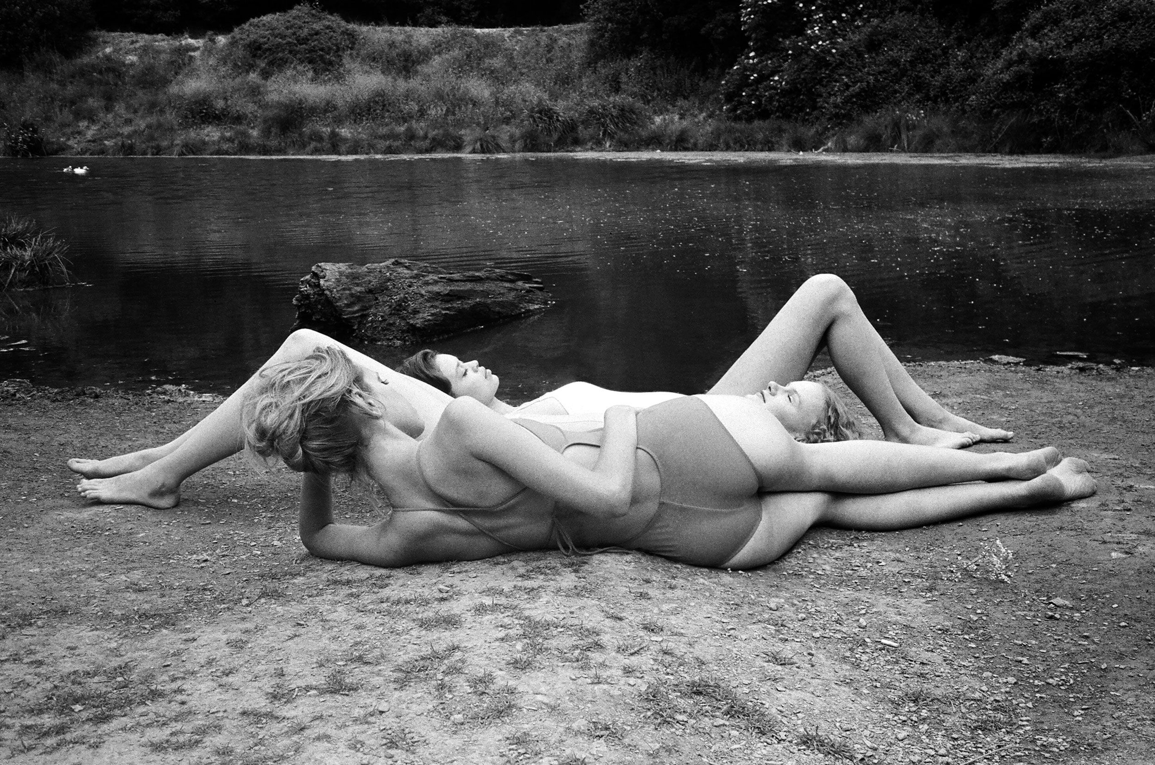 Three women in swimsuits are lying together on the ground near a body of water, trees, and vegetation in a black and white landscape.