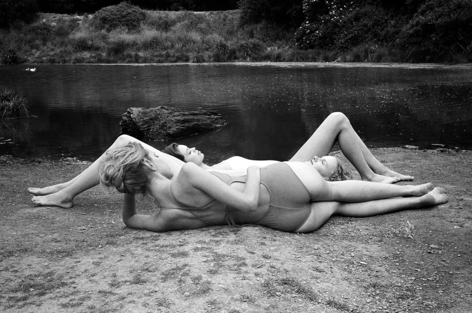 Three women in swimsuits are lying together on the ground near a body of water, trees, and vegetation in a black and white landscape.