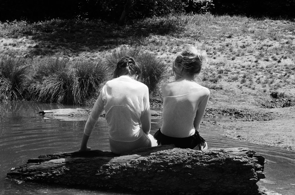 Black and white photo of two young women sitting side-by-side on a log in a pond, facing away from the camera, with grassy banks.