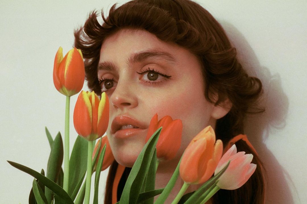 Close-up portrait of a young woman with dark hair and fair skin, surrounded by vibrant orange, yellow, and pink tulips in a soft indoor light.