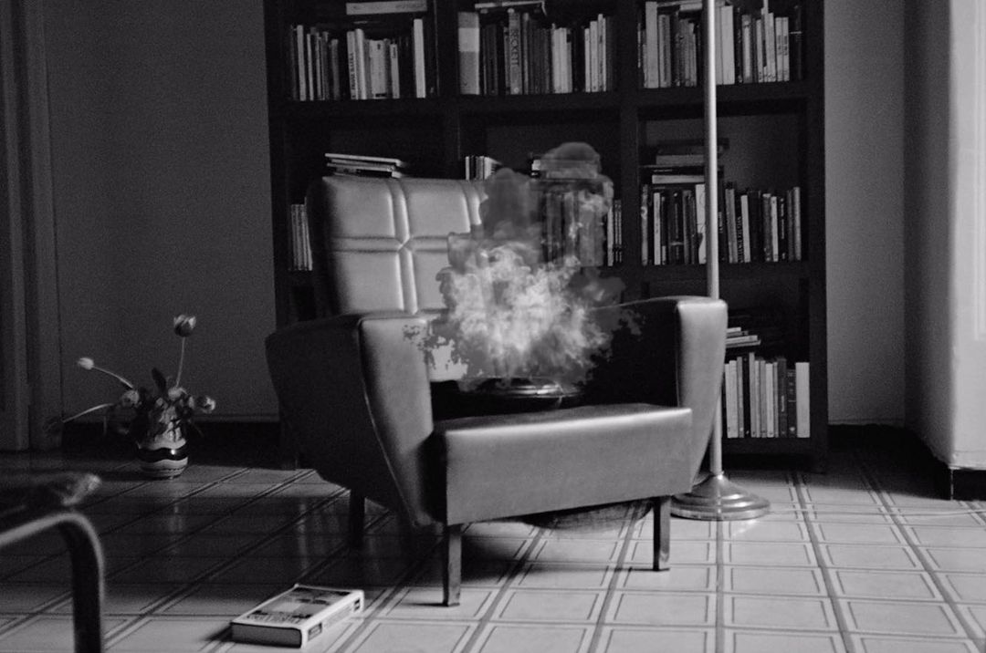 Black and white image of an armchair with smoke, a bookcase filled with books, and a vase with flowers in an indoor setting.