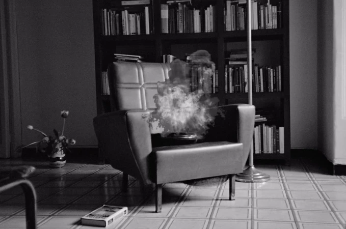 Black and white image of an armchair with smoke, a bookcase filled with books, and a vase with flowers in an indoor setting.