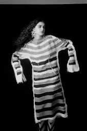 Woman with long curly hair wearing a striped tunic dress and large earrings against a dark background