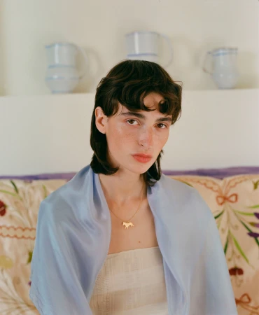 Portrait of a woman with short dark hair wearing a light blue silk scarf and gold necklace against a patterned sofa and ceramic jugs.