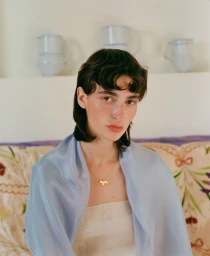 Portrait of a woman with short dark hair wearing a light blue silk scarf and gold necklace against a patterned sofa and ceramic jugs.