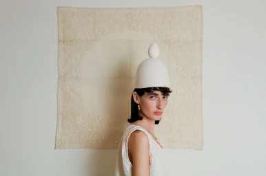 Portrait of a woman wearing a white sculptural dome hat with an egg-like top, positioned against an embroidered fabric hanging.