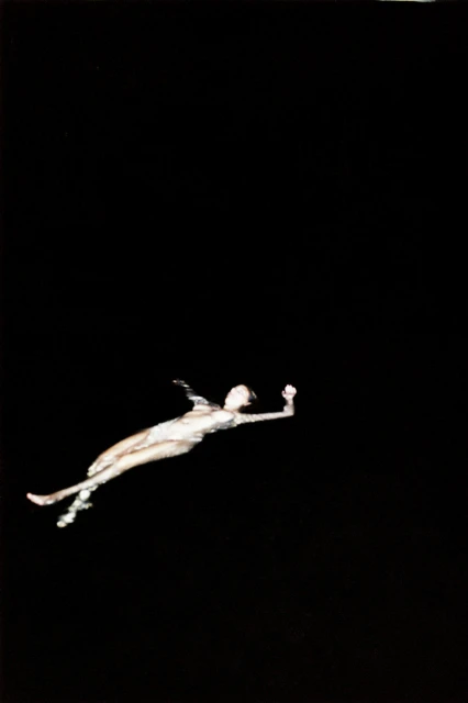 Artistic photograph of a female figure in an aquatic pose against a stark black background, creating a striking contrast and ethereal effect.