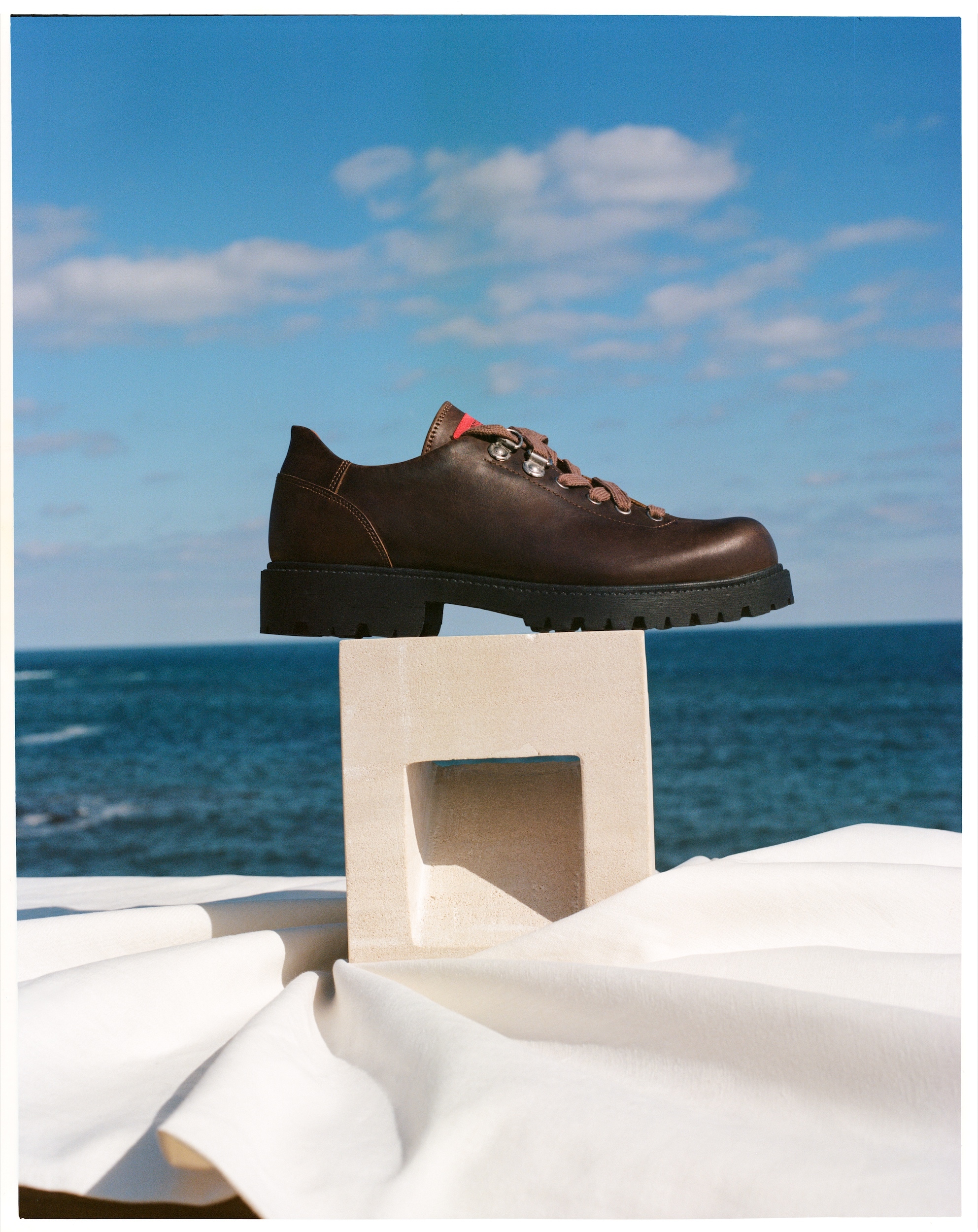 A dark brown leather shoe with tan laces sits atop a decorative white stand with a folded white cloth, with the ocean and sky behind it.