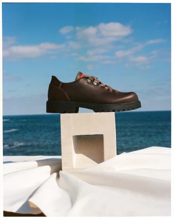 A dark brown leather shoe with tan laces sits atop a decorative white stand with a folded white cloth, with the ocean and sky behind it.