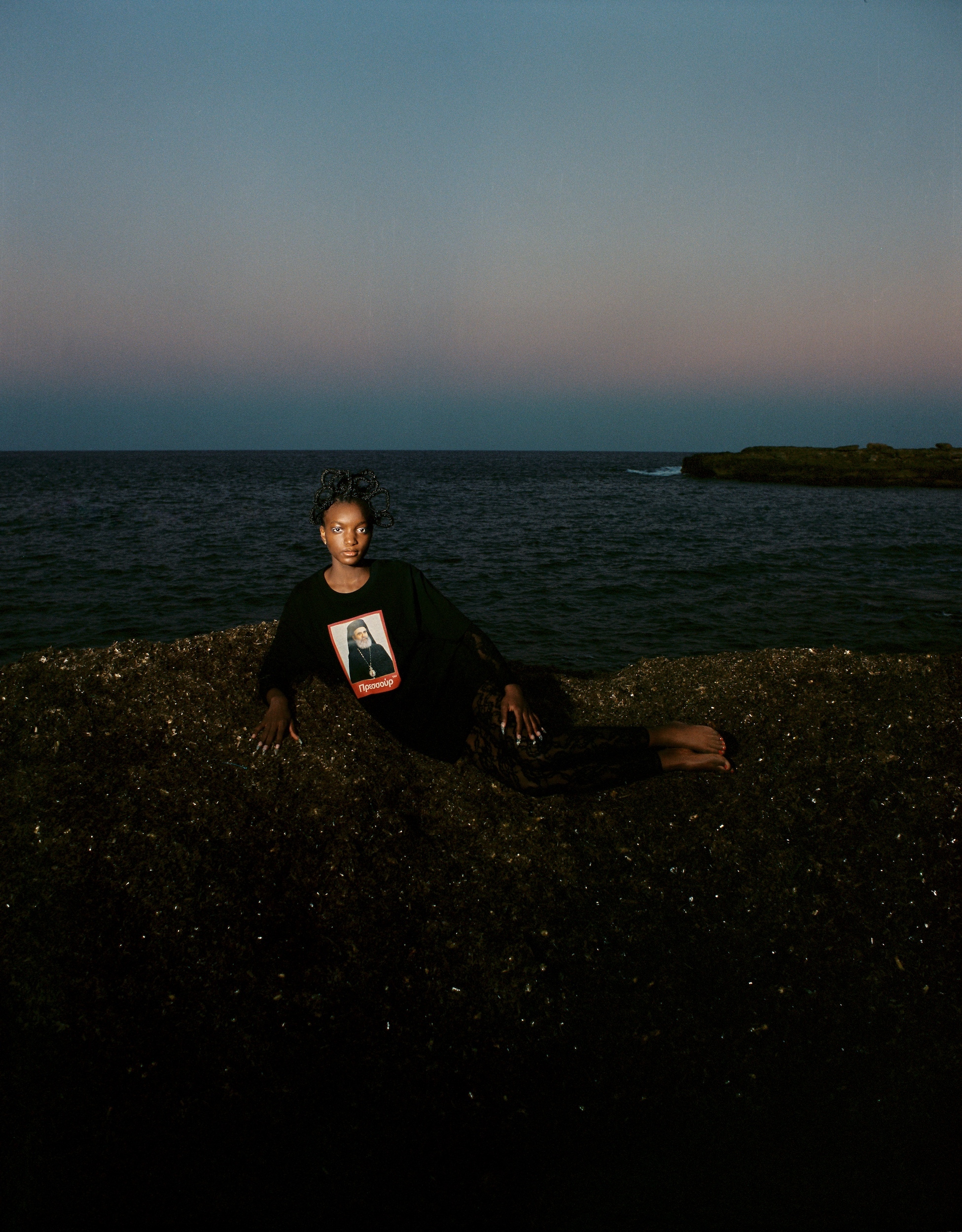 Woman in black long-sleeve shirt with graphic print lies on rocky beach at dusk with ocean and twilight sky visible in the background.
