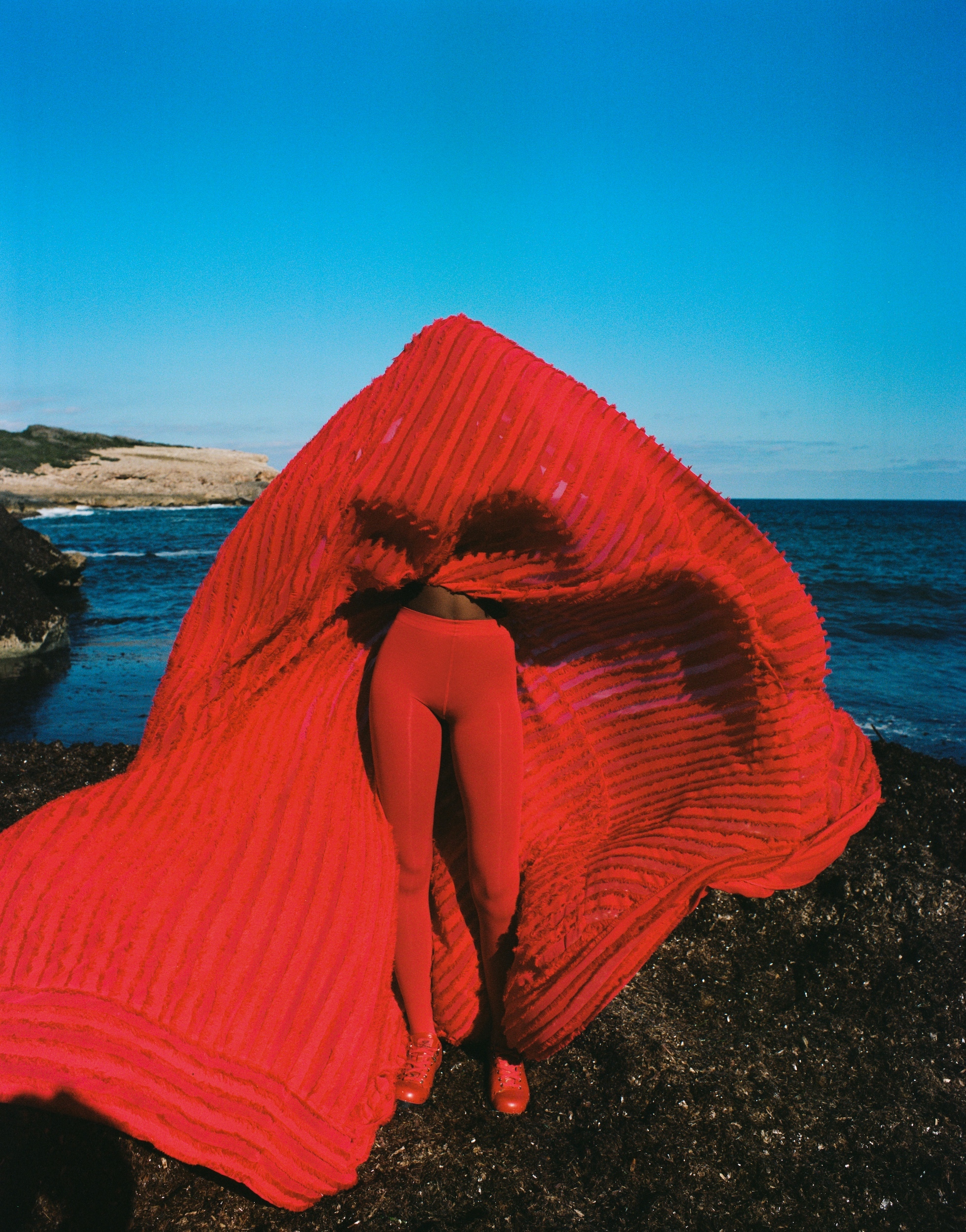 Person in a red outfit and matching draped fabric on a rocky coastline with ocean and blue sky backdrop, creating a striking visual.